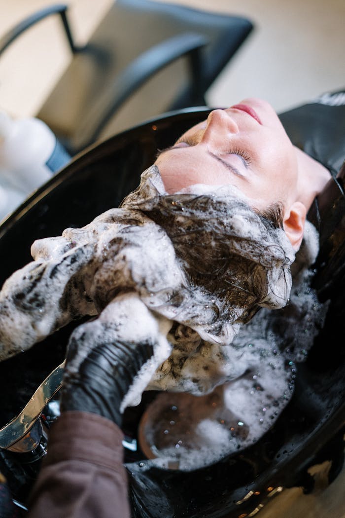 A woman getting her hair washed at a salon sink by a professional stylist wearing black gloves.